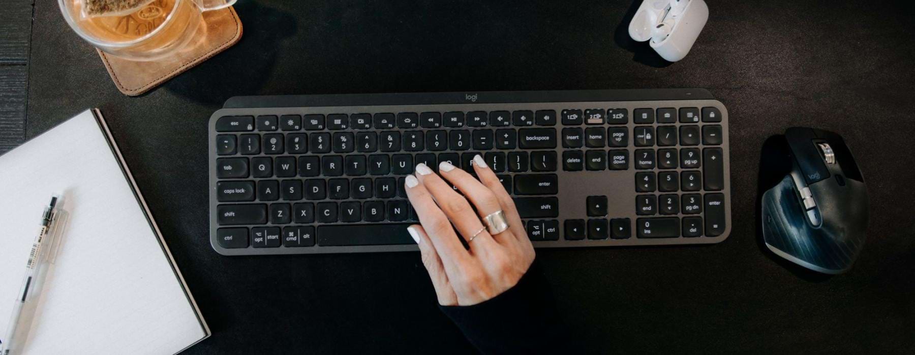woman's hand on a keyboard surrounded by office items and a cup of tea on a coaster